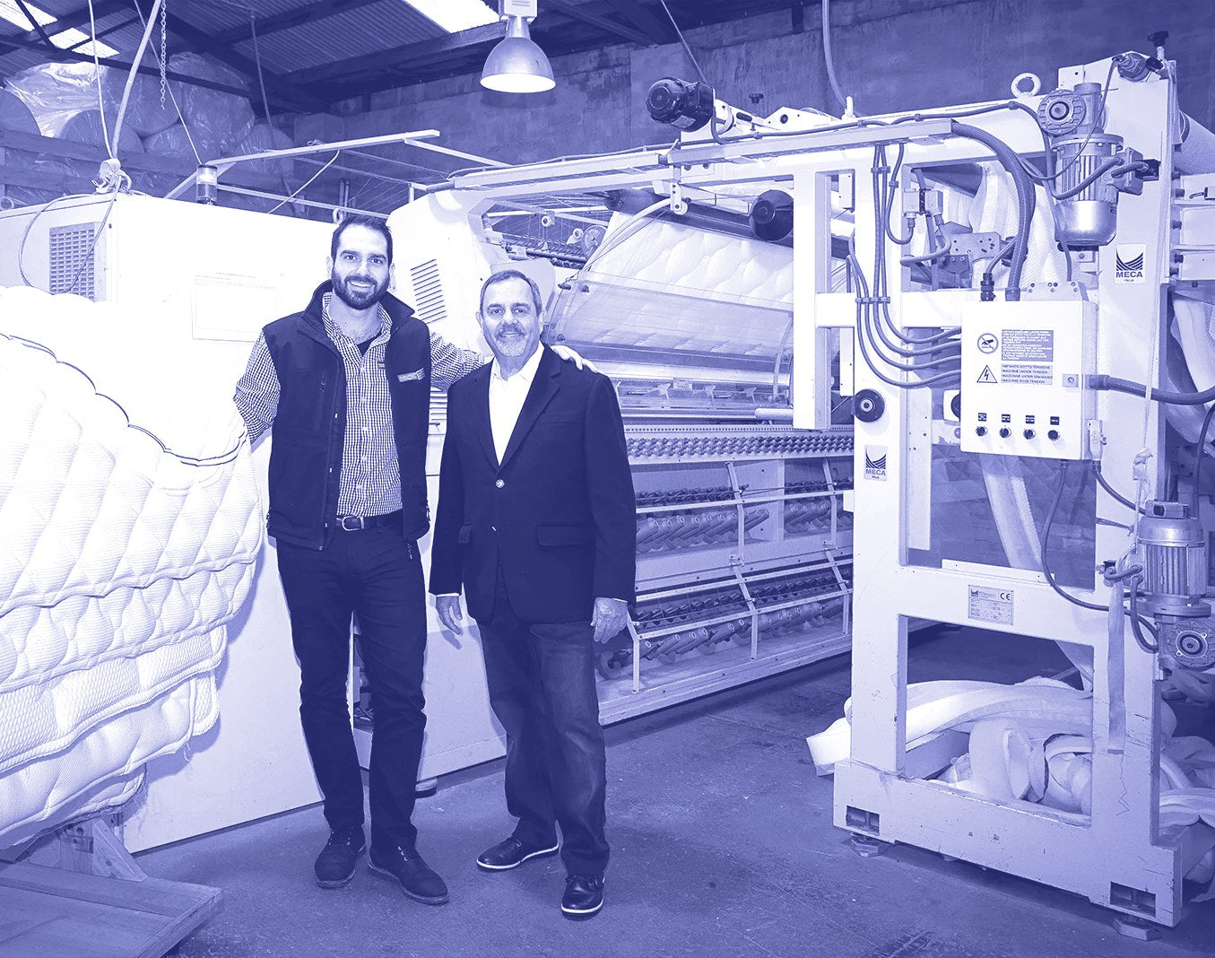 Two generations of the founding family standing beside the quilting machine on the factory floor